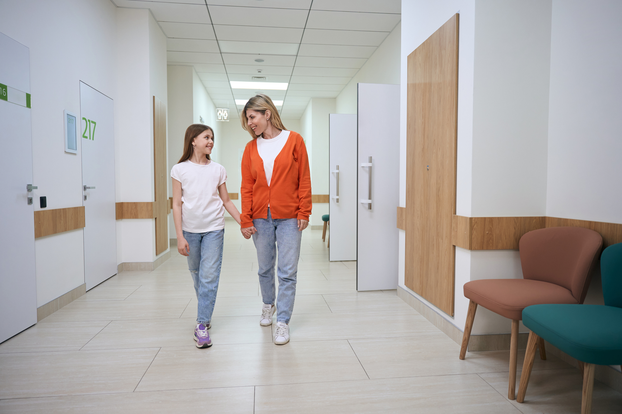 Woman in an orange sweater holds the girls hand, they walk along the hospital corridor.